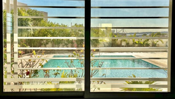 A view through a window with horizontal blinds, revealing a pool area with two swimming pools and surrounding greenery. - Gammarth Touristique Piscine Commune Bord de mer (La Marsa)