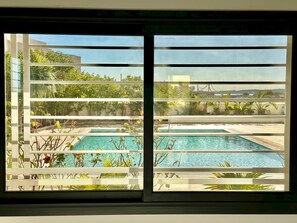 Une vue à travers une fenêtre équipée de stores horizontaux, dévoilant un espace piscine avec deux piscines et une végétation luxuriante environnante.