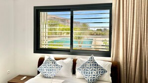 A hotel room with a bed, two pillows, a nightstand, a window with a view of a pool and greenery, and a curtain. - Gammarth Touristique Piscine Commune Bord de mer (La Marsa)