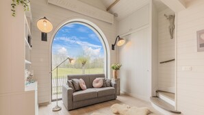 Una sala de estar acogedora con un sofá gris, un amplio ventanal de arco y una escalera de madera.