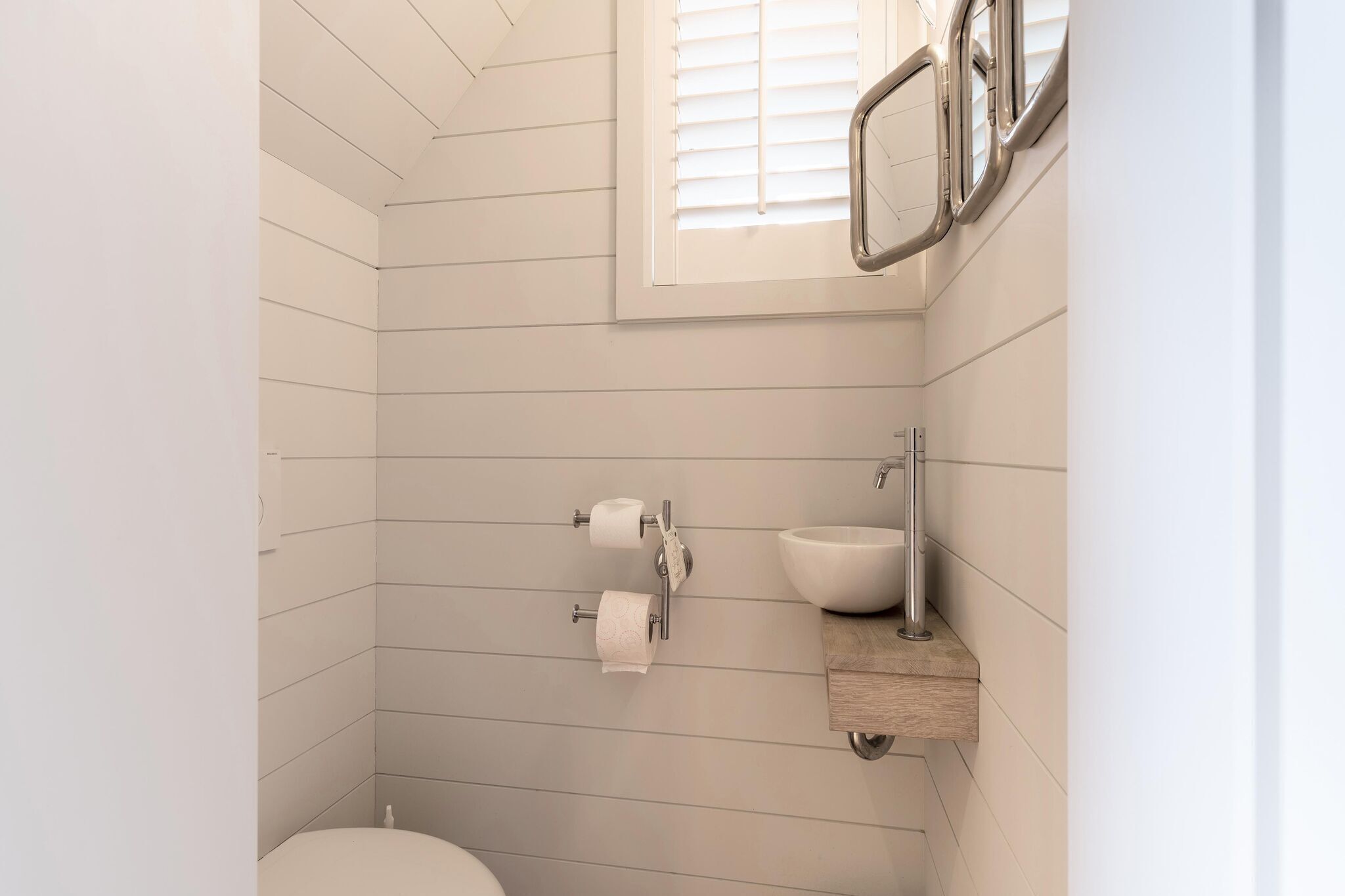 Ein Badezimmer mit weißen Fliesen, einer Toilette, einem Waschbecken und einem Fenster mit Jalousien.