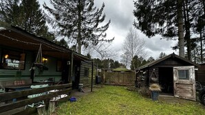 Een rustieke blokhut met een houten terras, een klein tuinhuisje en een gazon omringd door hoge bomen.