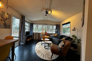 Ett mysigt vardagsrum med en soffa, fåtölj, soffbord och ett matbord. Det finns stora fönster med gardiner, en takfläkt och en vägglampa.