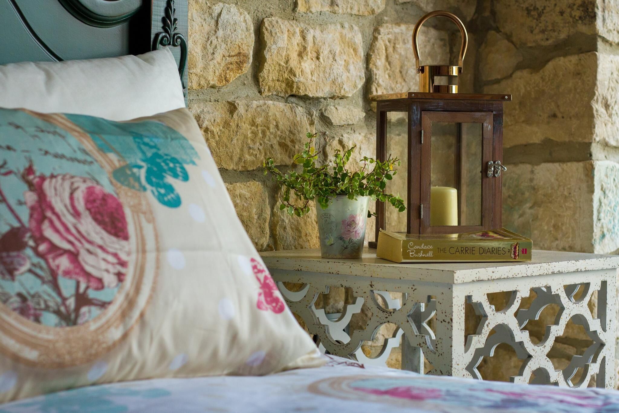 Una cama con un almohadón floral, una mesita de noche blanca pequeña con un farol, una planta en maceta y un libro titulado "The Carrie Diaries".