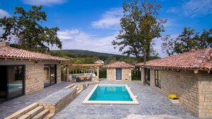 Uma área de piscina com paredes de pedra e telhados com azulejos, rodeada por árvores e um cenário montanhoso.