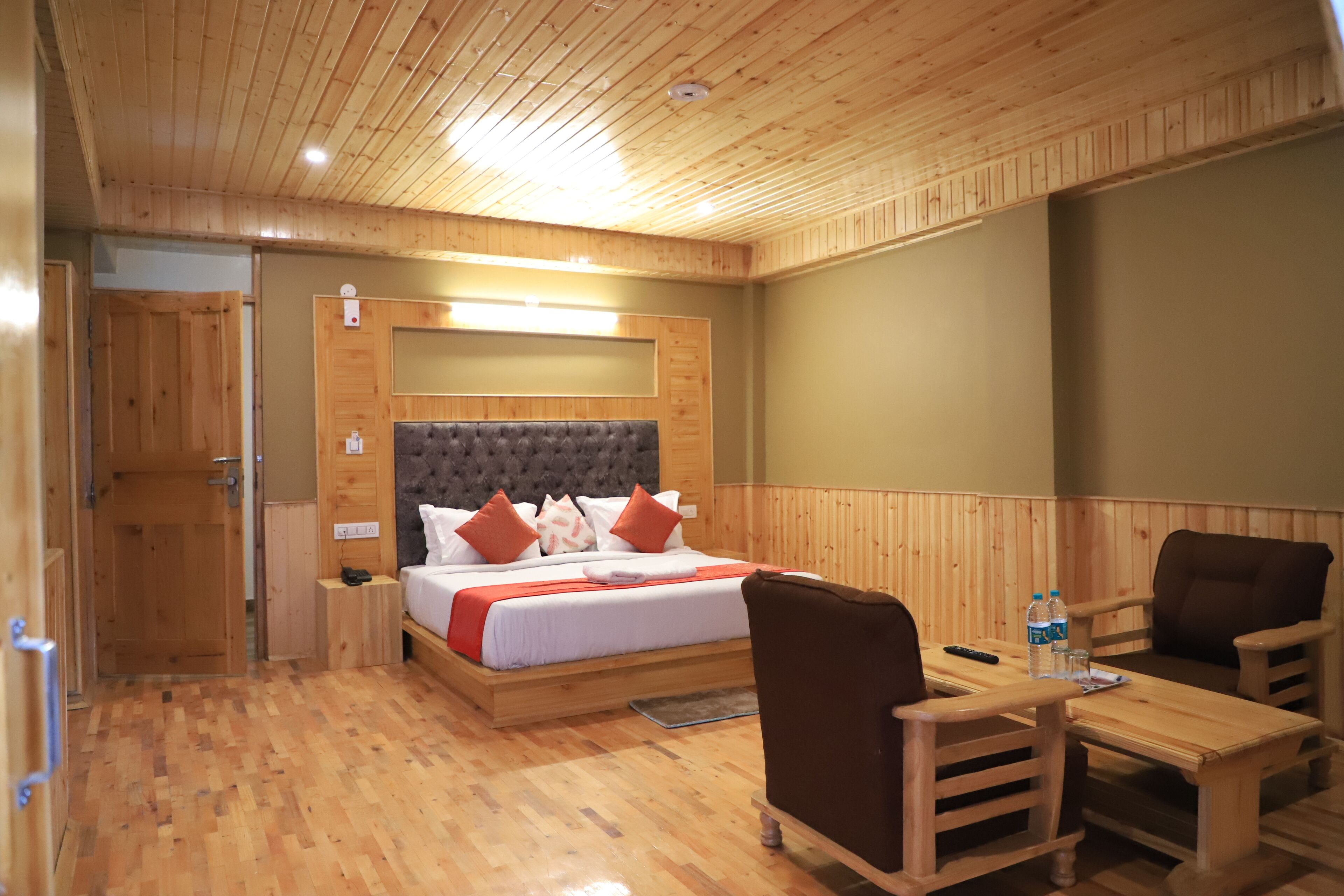 Une pièce au plancher de bois, avec un lit, deux chaises, une table et une porte en bois.