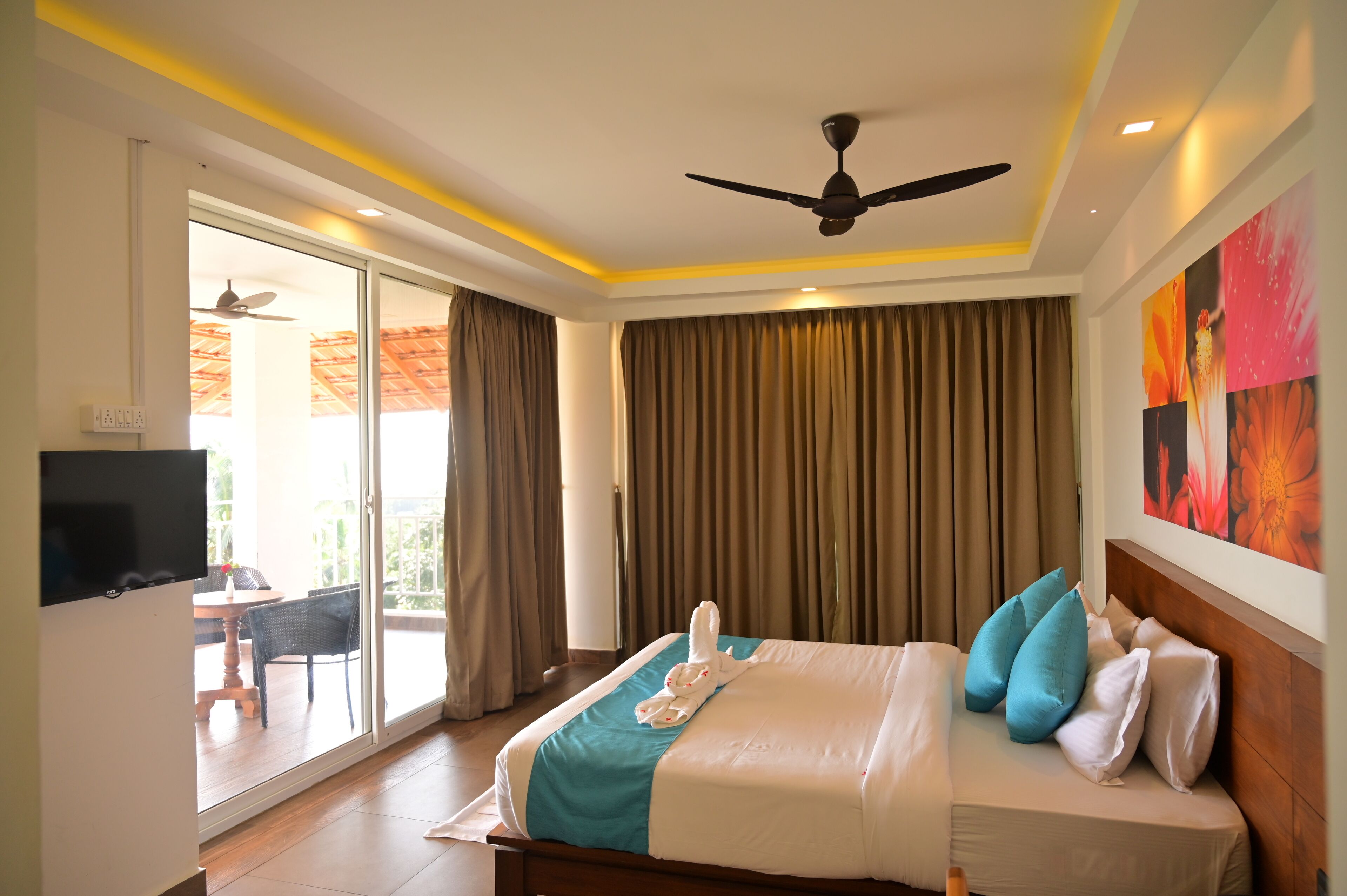 A hotel room with a bed, a ceiling fan, a TV, a balcony with a table and chairs, and a painting on the wall.