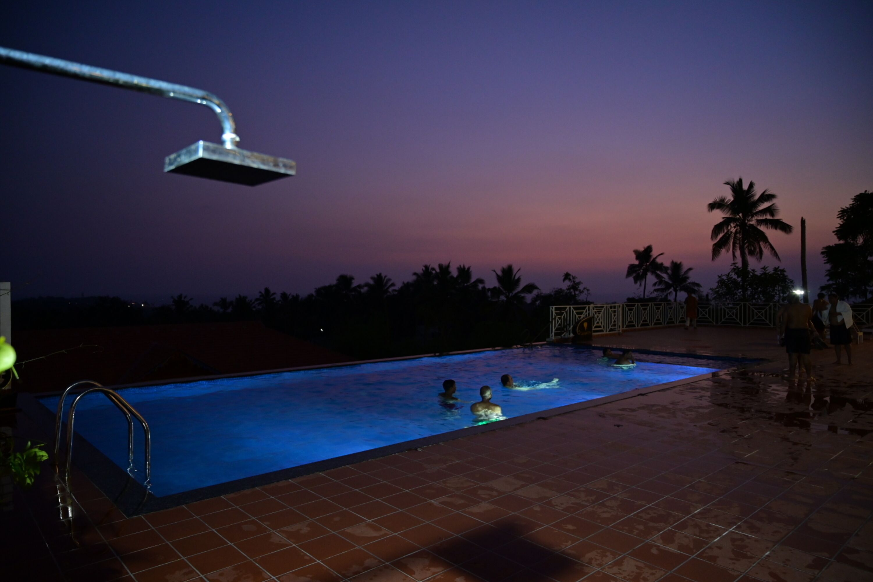 Una piscina al anochecer con vista al atardecer, un rociador y un deck con baldosas.