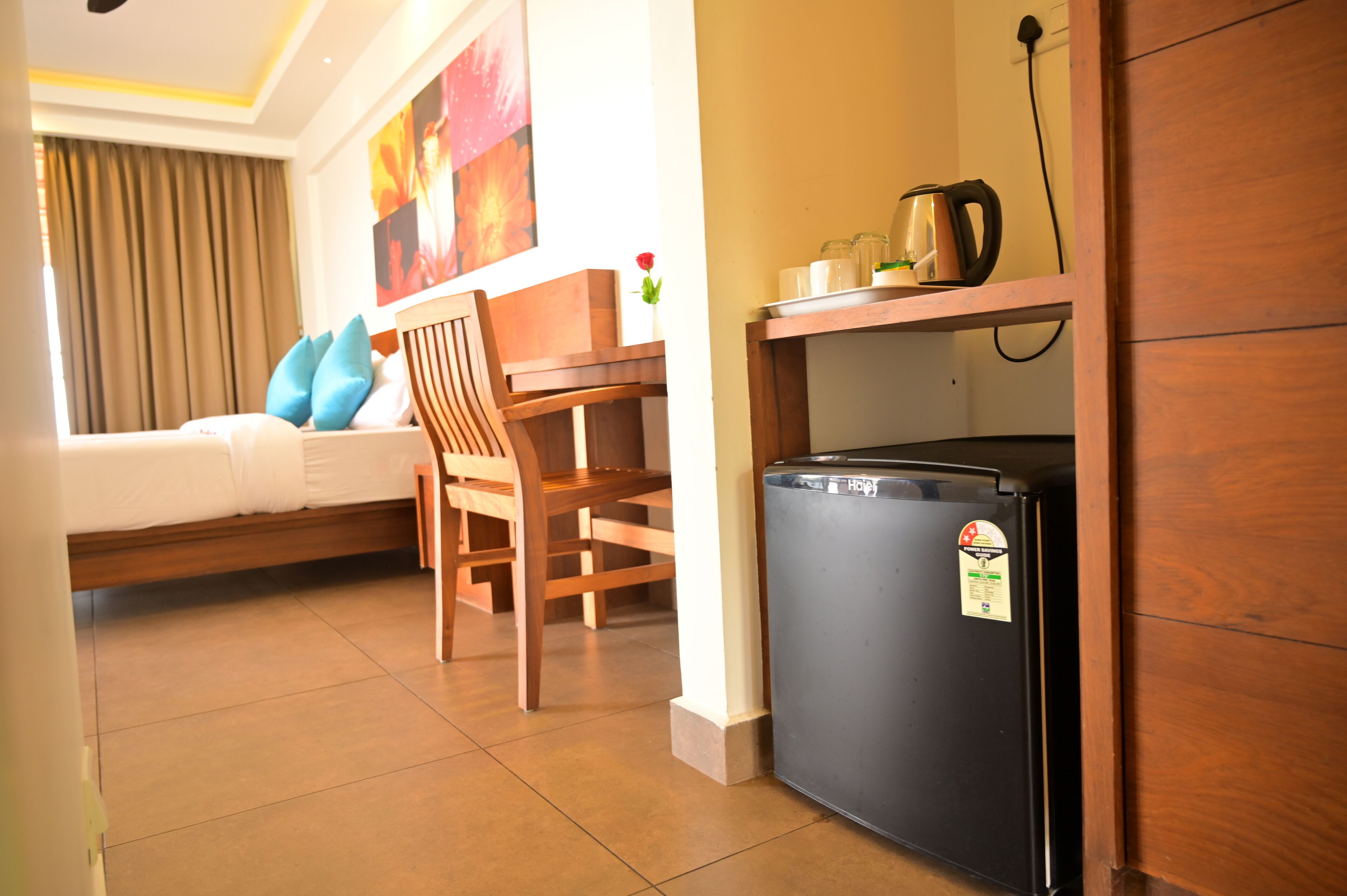 A compact hotel room with a bed, a wooden chair, a small table, a black refrigerator, and a wall-mounted shelf with a kettle and cups.