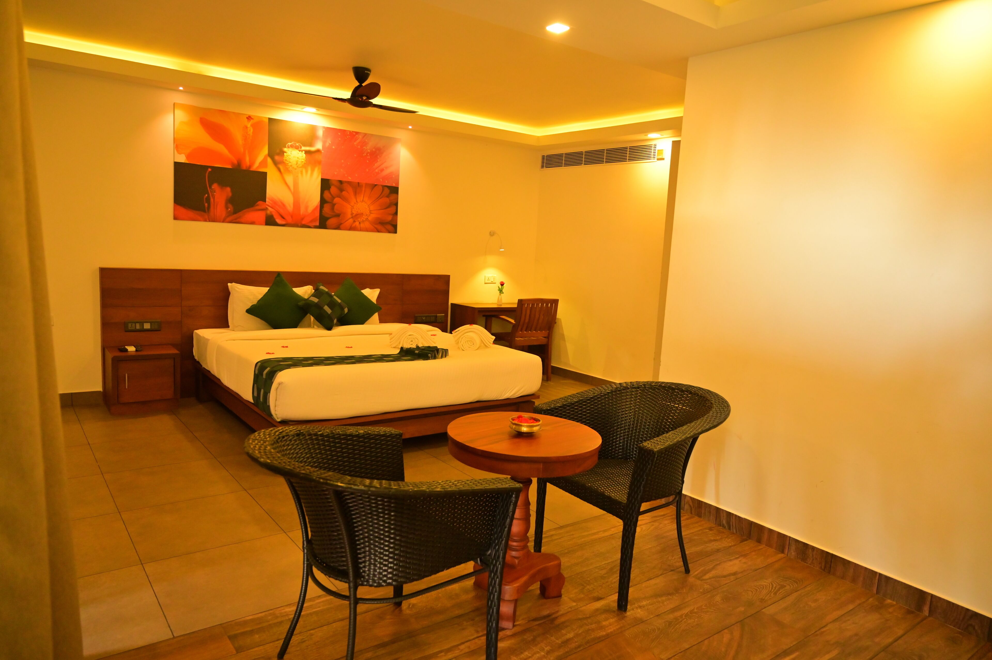A hotel room with a bed, two chairs, a small table, a ceiling fan, and artwork on the wall.