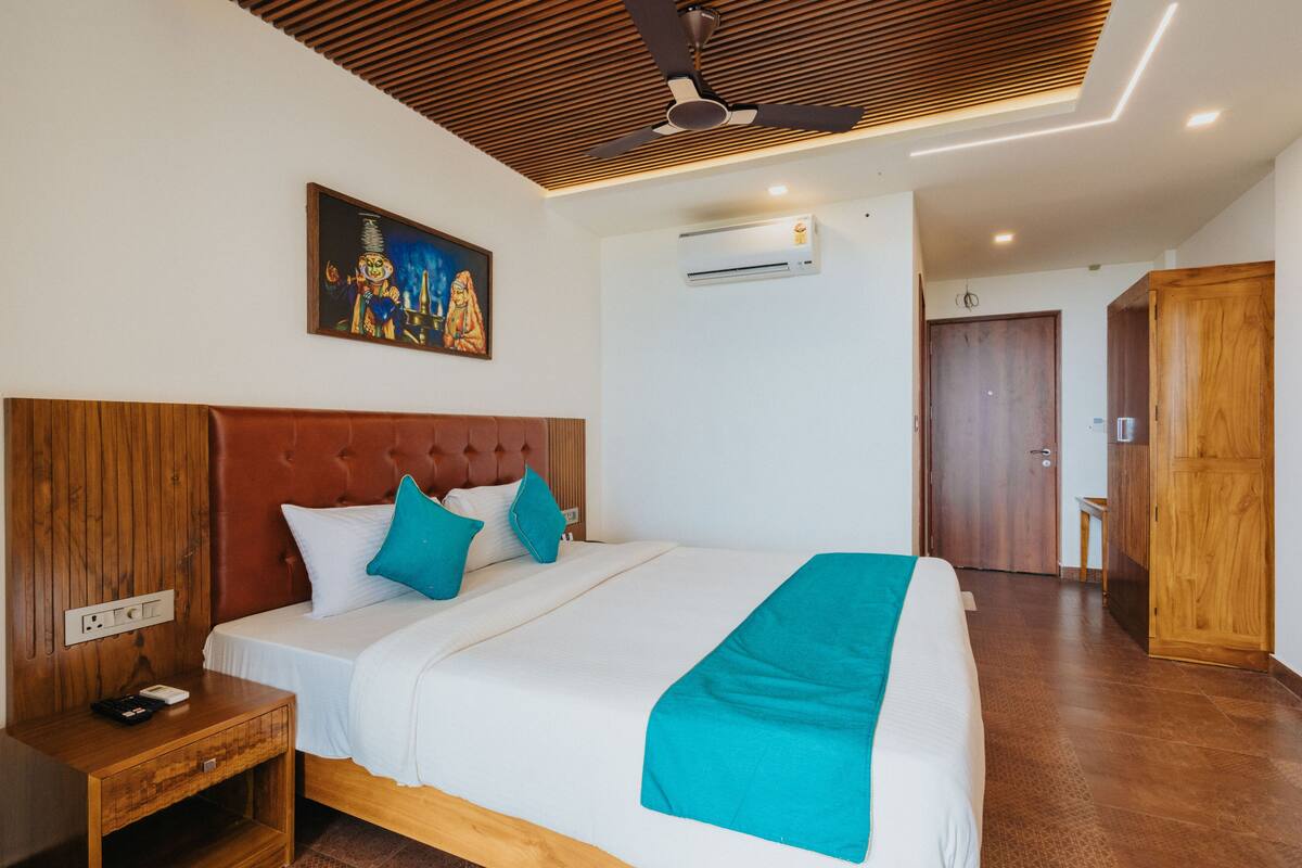 A hotel room with a large bed, wooden headboard, a ceiling fan, an air conditioning unit, and a painting on the wall.