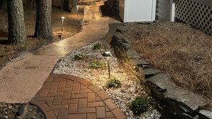 Un chemin extérieur illuminé, bordé de guirlandes lumineuses, qui mène à une maison entourée d’arbres et d’un mur de pierres.