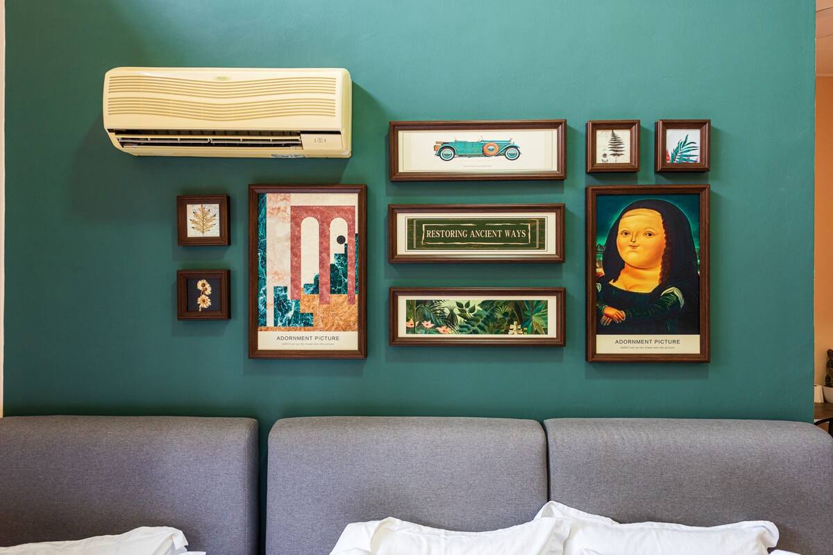 A room with a teal wall, air conditioning unit, and a series of framed pictures including 'Restoring Ancient Ways' and 'Adornment Picture'.