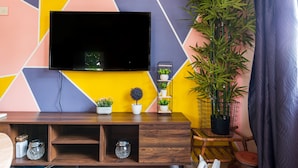 En moderne stue med et fladskærms-tv monteret på en væg med et farverigt geometrisk mønster, et træunderholdningsmøbel med dekorative genstande, og en stor potteplante.