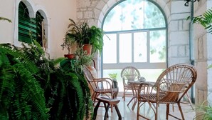 Una habitación con pared de piedra, un amplio ventanal arqueado, mobiliario de mimbre y exuberante vegetación.