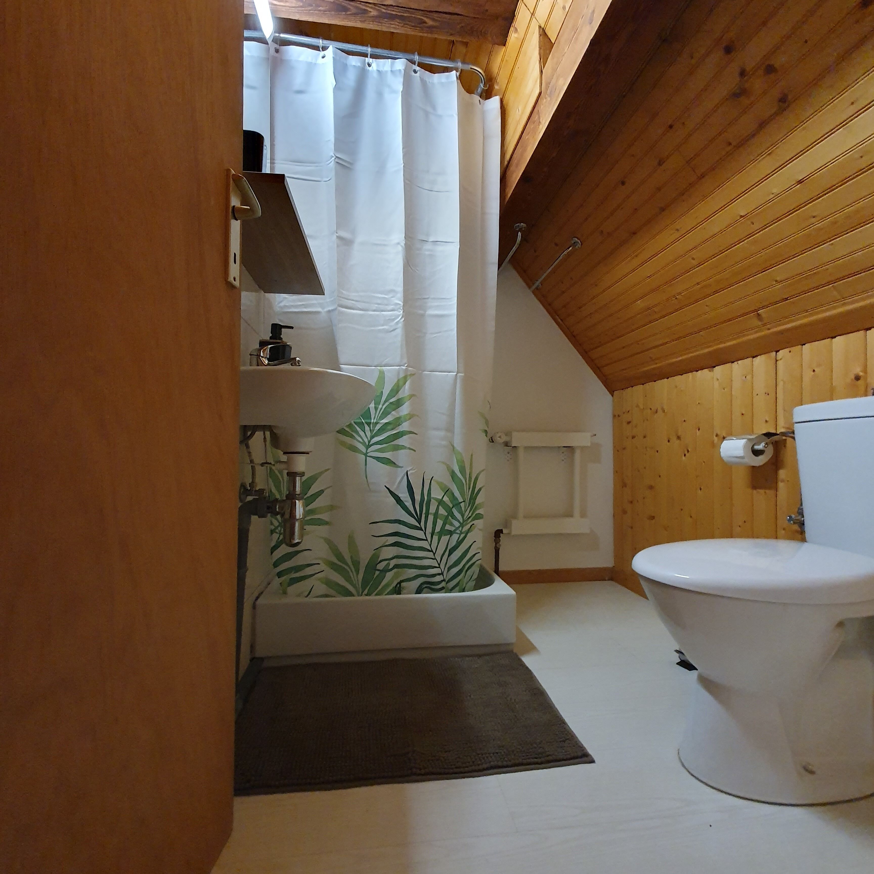 Une petite salle de bain aux murs lambrissés, avec des toilettes blanches, une douche dotée d’un rideau blanc et un lavabo avec un robinet.
