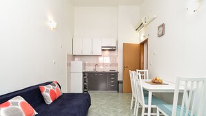 A compact living space with a dark blue sofa, a white dining table with chairs, a kitchen area with cabinets and a refrigerator, and a wooden door.