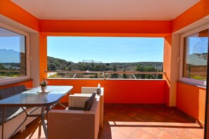 Ein Balkon mit orangefarbenen Wänden, einem Glastisch, Korbstühlen und Blick auf eine hügelige Landschaft.