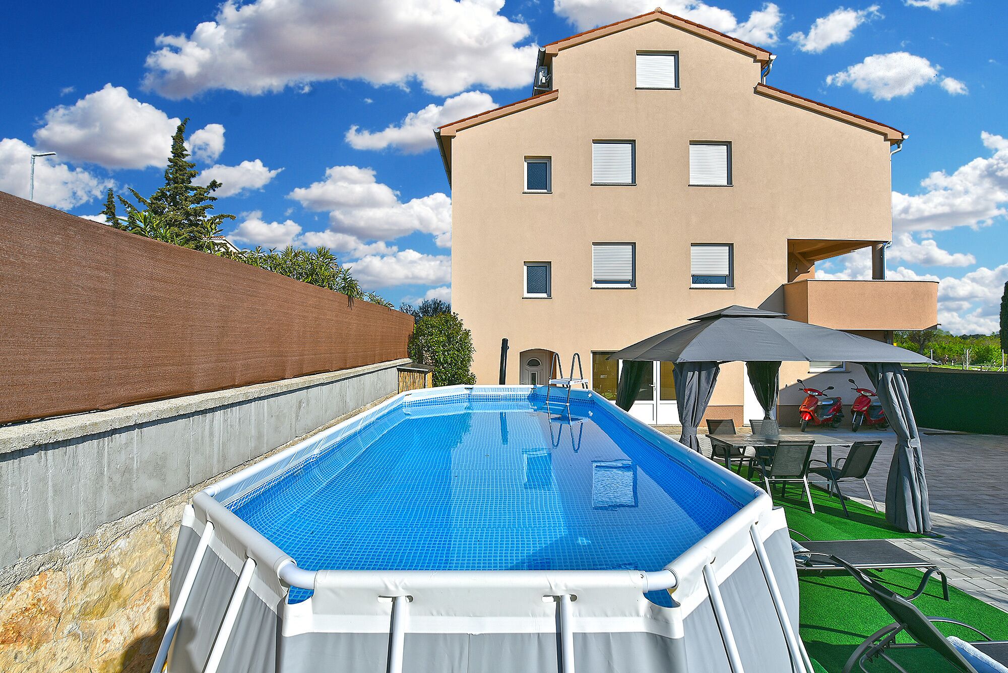 A swimming pool with a ladder, a covered seating area, and a building in the background.