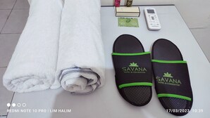 Toalha branca dobrada, um par de chinelos pretos com detalhes verdes, um pequeno aparelho branco e dois frascos de shampoo sobre uma superfície branca.