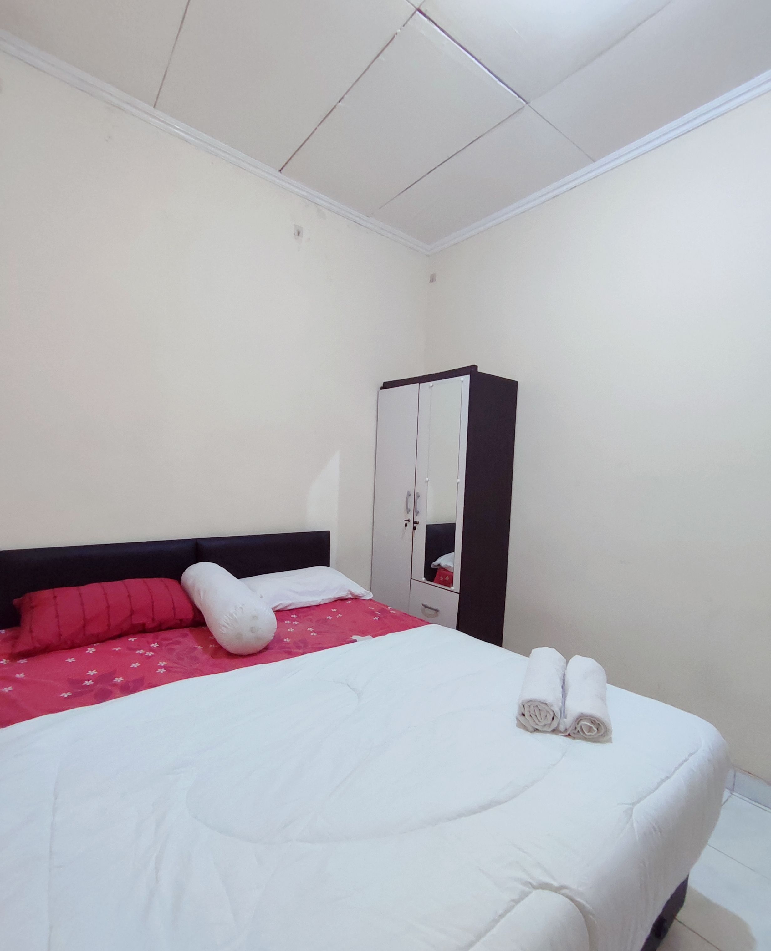 Un lit bien fait, avec une literie blanche et rouge, une tête de lit sombre, une armoire avec un miroir et une petite serviette pliée posée sur le lit.