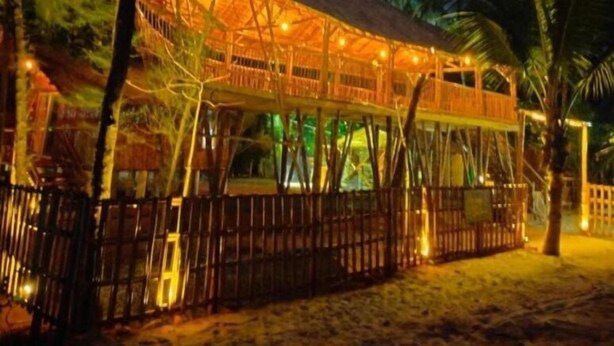 A wooden structure with a roof, surrounded by trees and palm trees, likely a resort or lodge.