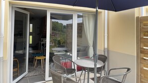 Eine Glastür führt zu einer Terrasse im Freien mit Tisch und Stühlen, einem dunklen Sonnenschirm und Blick auf die Umgebung.