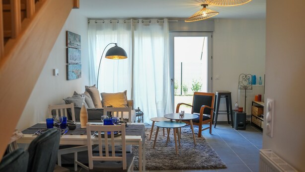 Ein modernes Wohnzimmer mit Sofa, Esstisch, Stühlen, einer Treppe und einem großen Fenster mit Vorhängen.