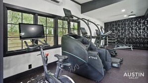 A gym with treadmills, an elliptical, and a stationary bike. There is a TV on a stand and a wall with geometric patterns. - Amenity Heavy Serene Apt in Convenient Location (Austin)