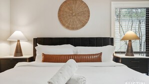 A hotel room with a large bed, two bedside tables with lamps, a round wall decoration, and a window with blinds. - Amenity Heavy Serene Apt in Convenient Location (Austin)