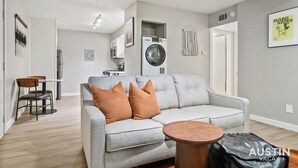 A modern living room with a grey sofa, a round coffee table, and a washing machine in the background. - Amenity Heavy Serene Apt in Convenient Location (Austin)