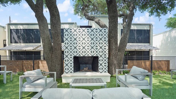 A modern outdoor patio with a built-in fireplace, surrounded by white furniture and large trees. - Amenity Heavy Serene Apt in Convenient Location (Austin)