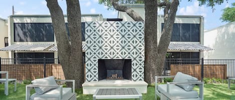 A modern outdoor patio with a built-in fireplace, surrounded by white furniture and large trees.