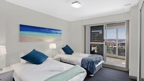 A hotel room with two beds, a painting of a beach, a window with blinds, and a view of buildings.