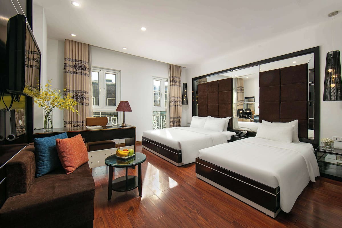 Quarto de hotel moderno com duas camas, um sofá, uma mesa pequena com frutas e um espelho grande.