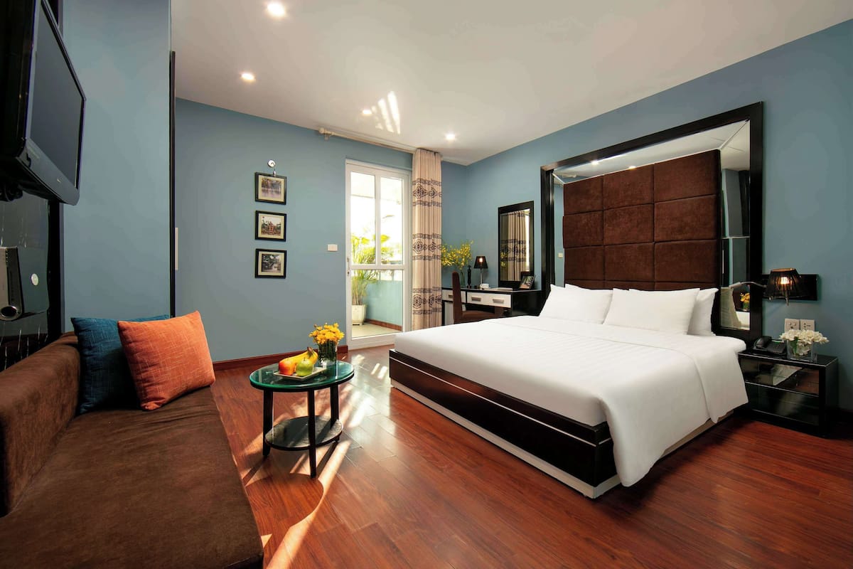 Quarto de hotel moderno com uma cama grande, um sofá, uma mesinha com flores, uma TV e um espelho amplo.