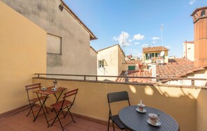 A balcony with a table and chairs, overlooking a cityscape with terracotta roofs.