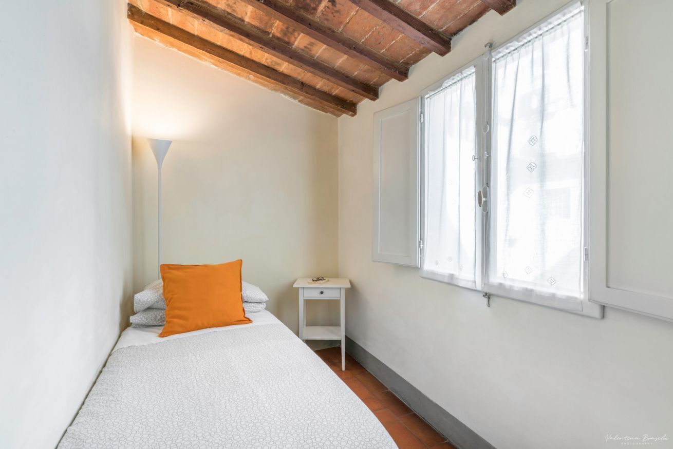A small, bright room with a bed, a nightstand, and a window.