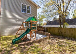 Playful backyard oasis with a wooden play structure and green slide, surrounded by a tree and a wooden fence.