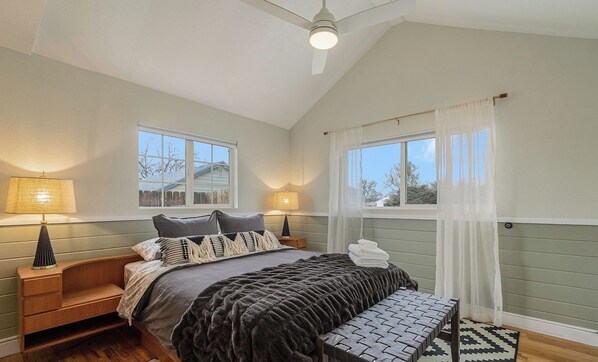 A bedroom with a bed, bedside tables, a bench, and two windows with curtains.