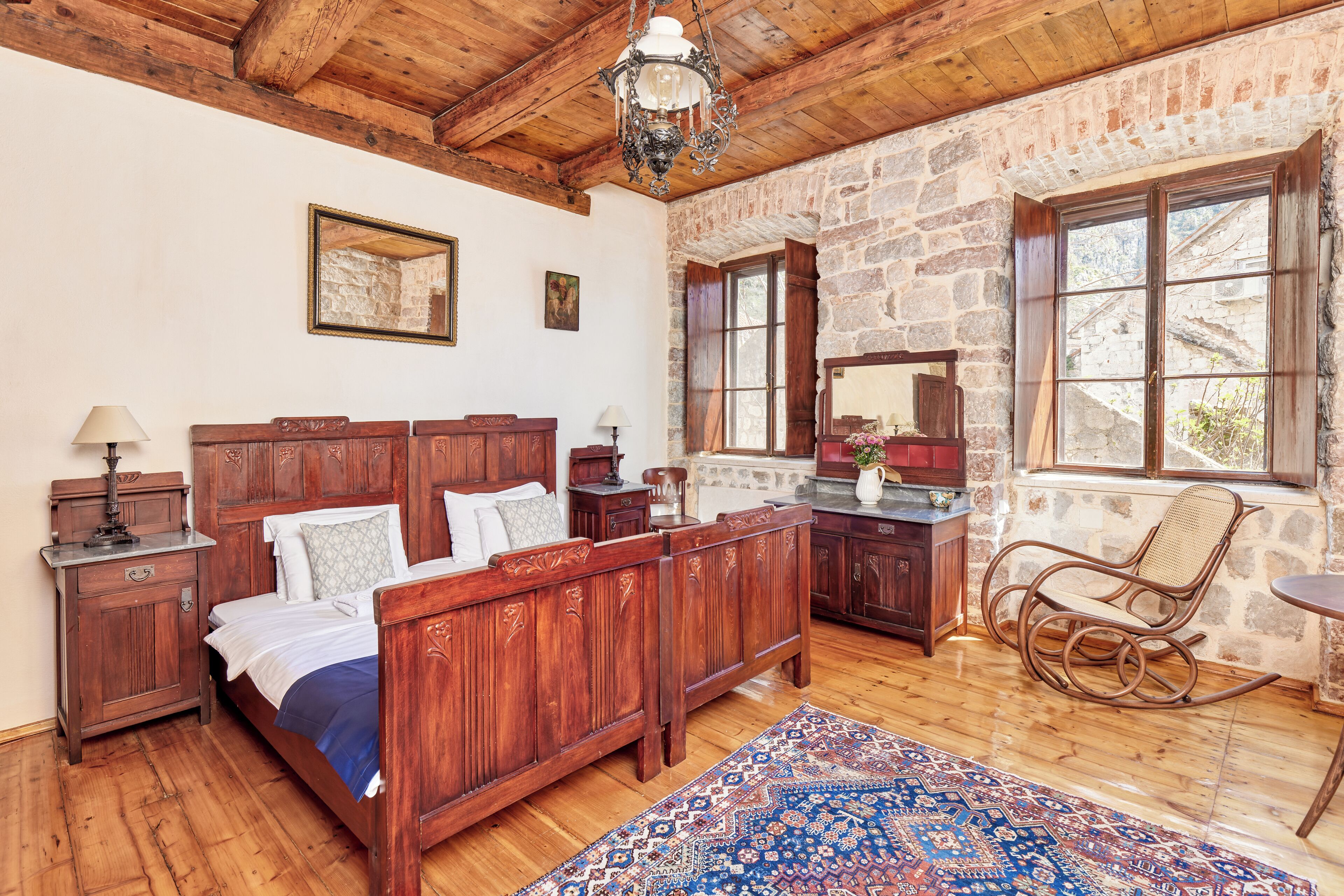 Una cama de madera con cabecera, una mesita de noche, un sillón mecedora y una pared de piedra.