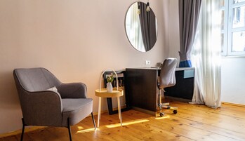 Una habitación moderna con un sillón gris, una mesita de noche con un florero, un escritorio con una silla, un espejo redondo y una ventana con cortinas.