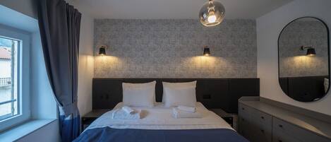 Una habitación de hotel moderna con una cama grande, dos lámparas fijadas a la pared, un espejo redondo y una ventana con cortinas.