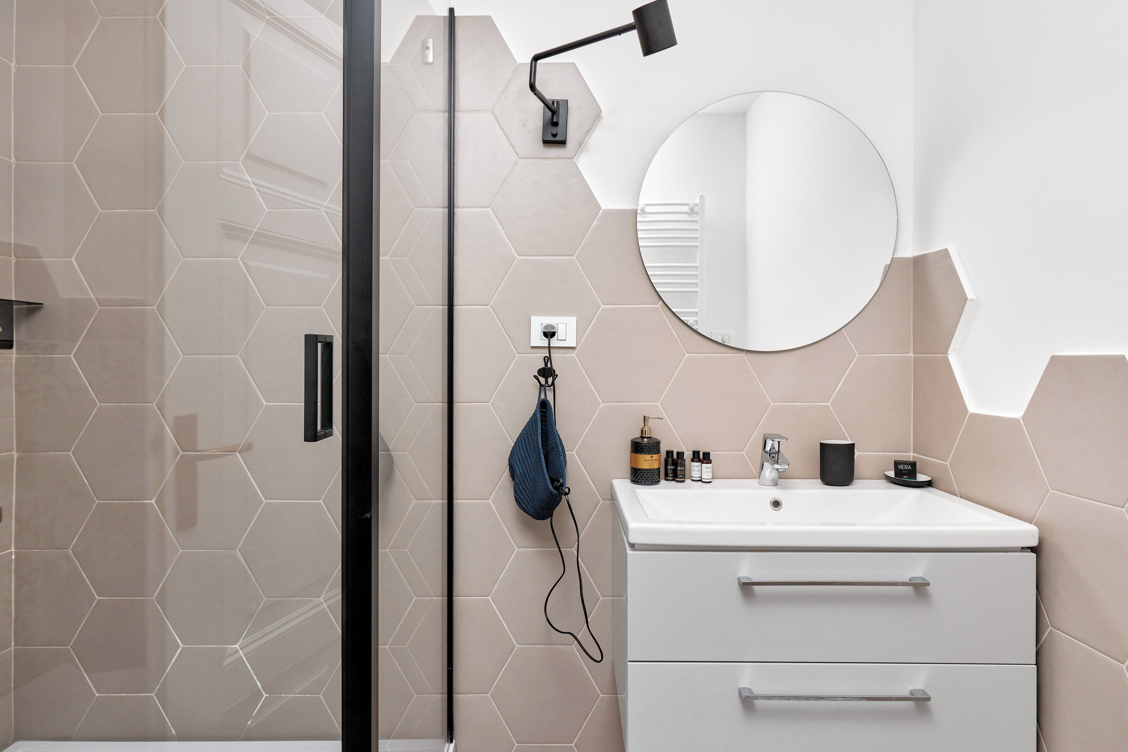 A bathroom with a hexagonal tile wall, a round mirror, a white sink with a towel, and a glass shower enclosure.