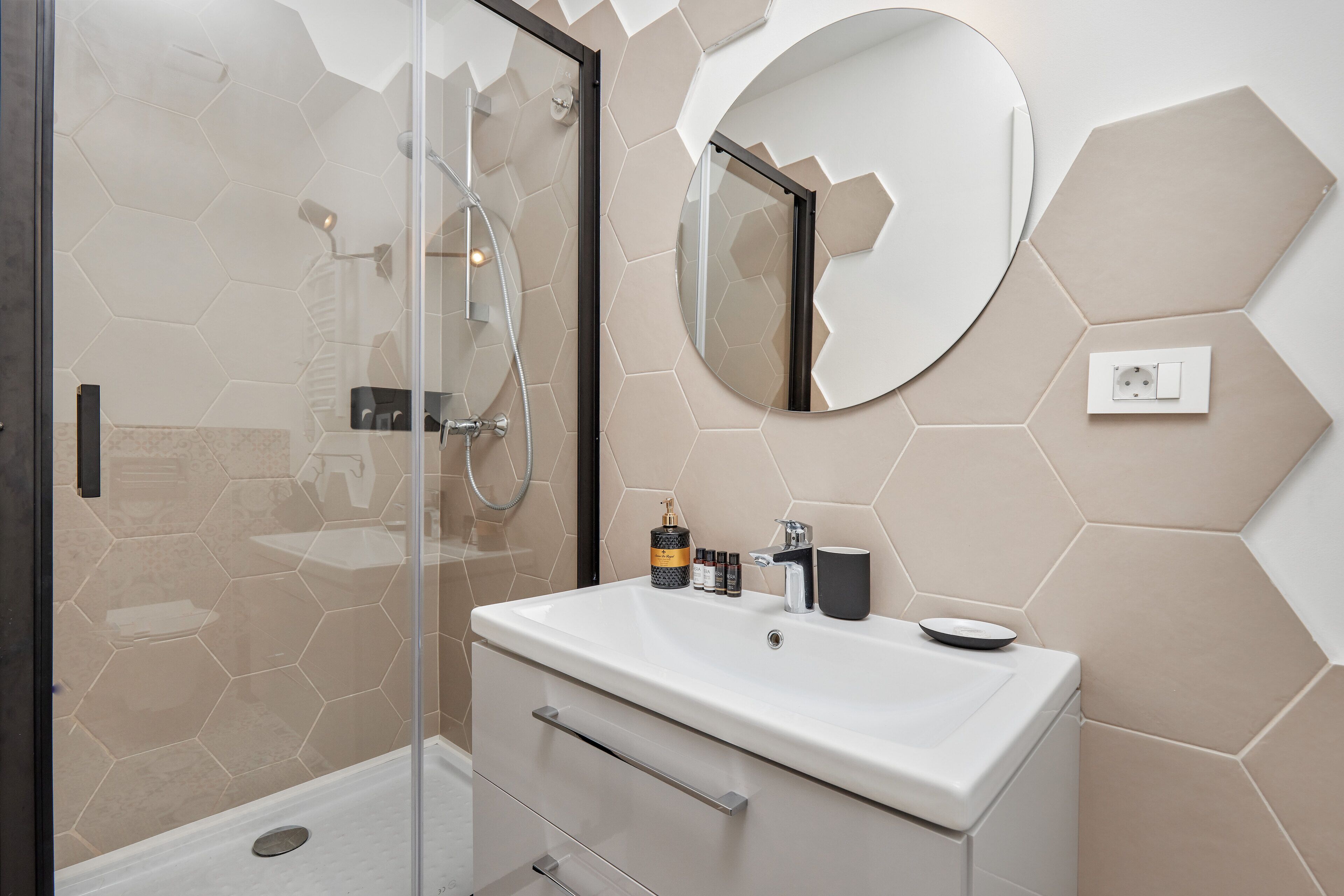 Un baño moderno con ducha envuelta en vidrio, un espejo redondo y un lavabo blanco con artículos de tocador.