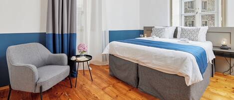 Habitación de hotel con cama, una silla, una mesita con un florero y ventana con vistas a edificios.