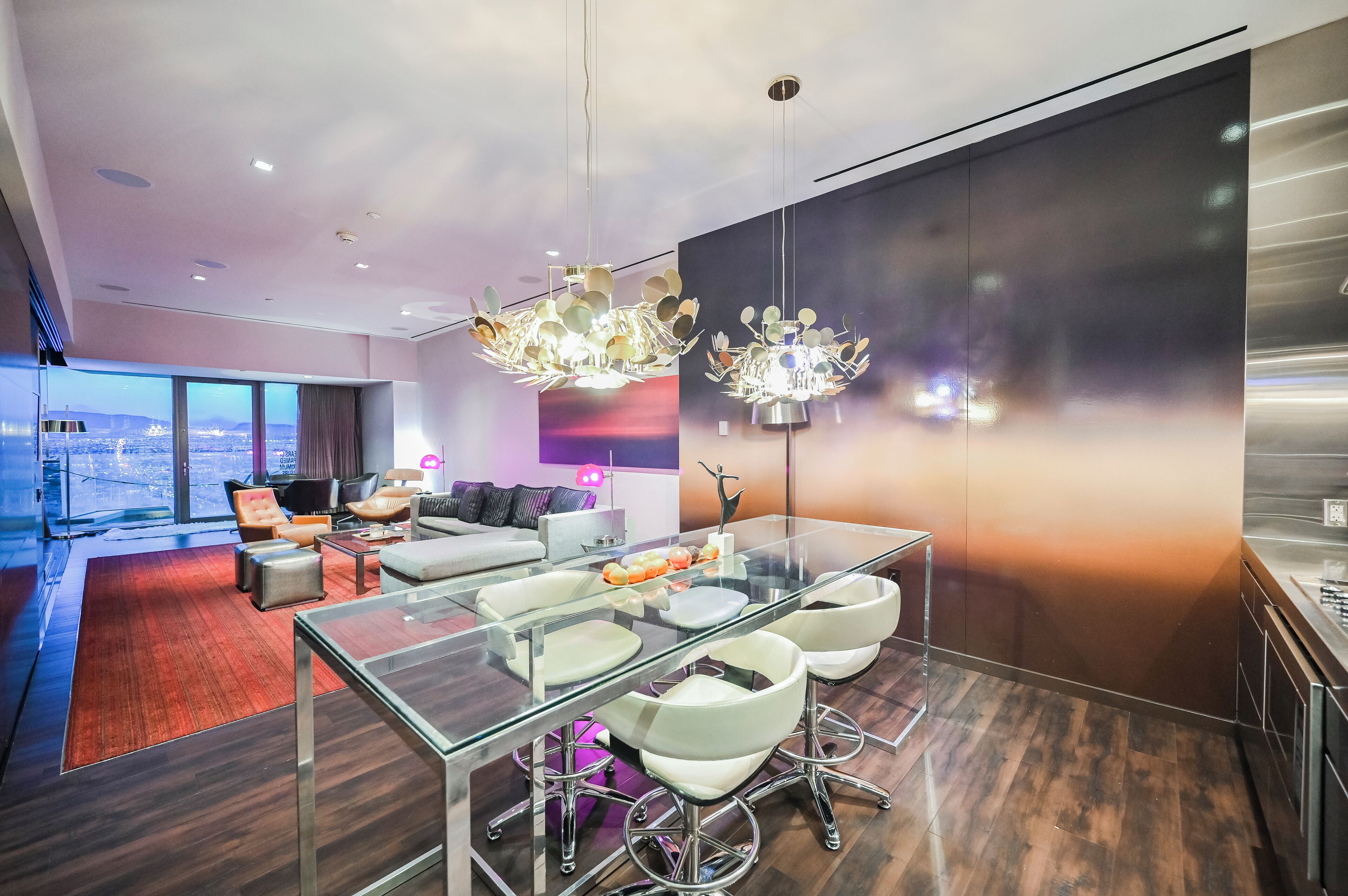 Una habitación de hotel moderna con una mesa de comedor de vidrio, sillas blancas y vistas a la ciudad.