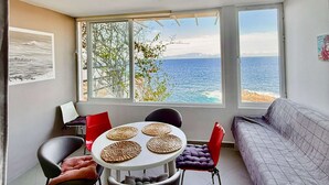 Ein Zimmer mit großem Fenster mit Meerblick, einem runden Tisch mit gewebten Untersetzern, einem grauen Sofa und einem roten Sessel.