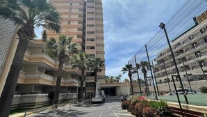 Un edificio de varios pisos con una entrada central, rodeado de palmeras y una cancha de tenis cercada.