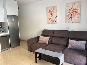 A modern kitchen with a built-in refrigerator, a dining area with a bench, and a living room with a sofa and two floral paintings on the wall.